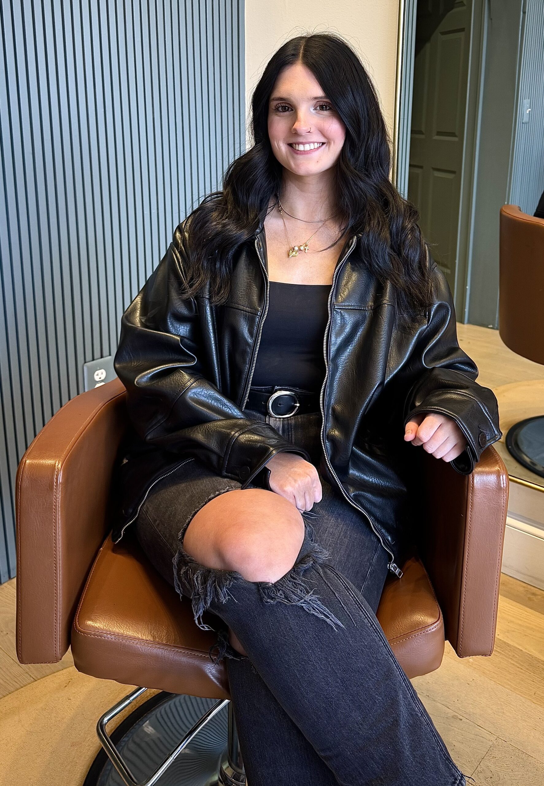 Smiling young woman with long dark hair wearing a black leather jacket and ripped jeans, seated in a brown salon chair.