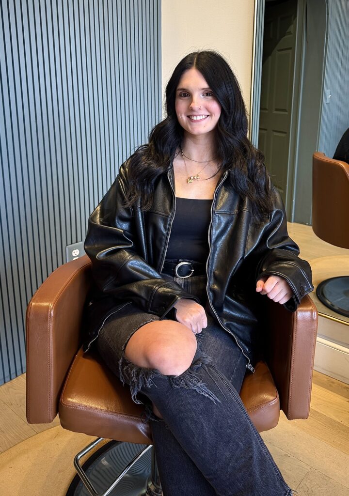 Young woman with long dark hair wearing a black leather jacket and ripped jeans sits in a brown salon chair, smiling at the camera.